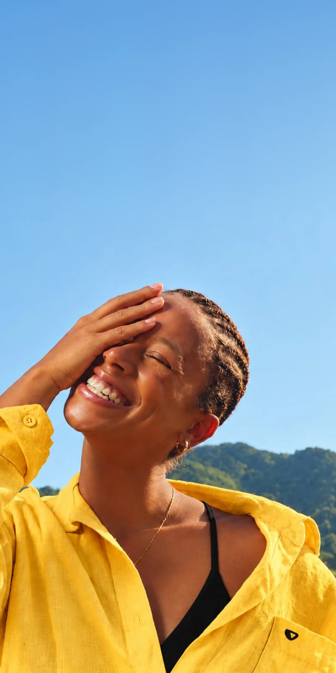 A young woman in a yellow shirt laughing in the sun, face turned up to an open blue sky